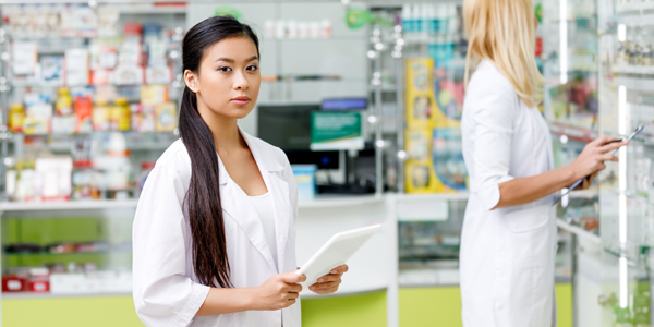 A pharmacist wearing a white coat stands behind the counter in a pharmacy with green walls, organizing medication bottles. Online Prescription Transfer Form